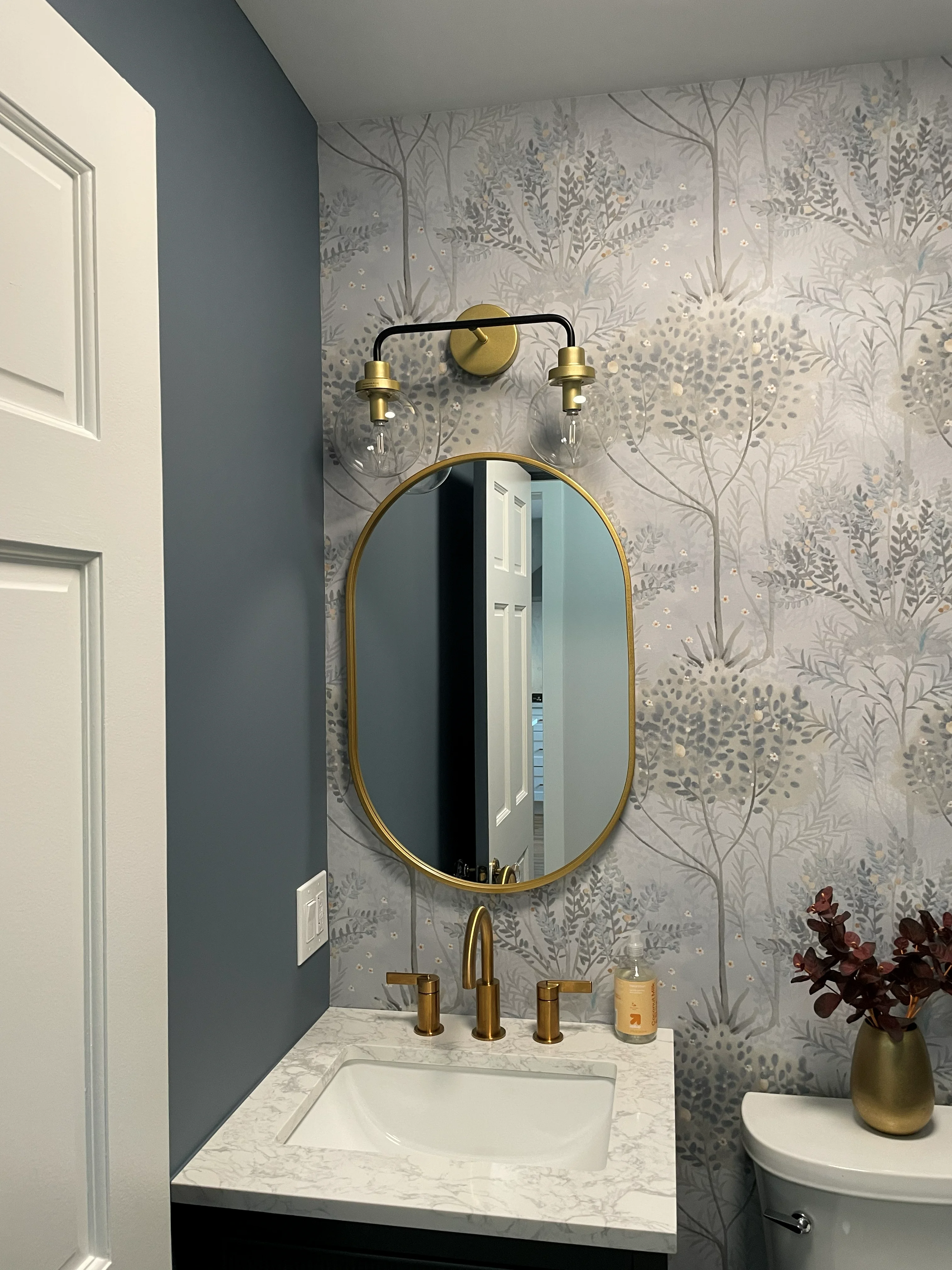 Marble vanity with brass mirror, fixtures, and botanical wallpaper in modern bathroom