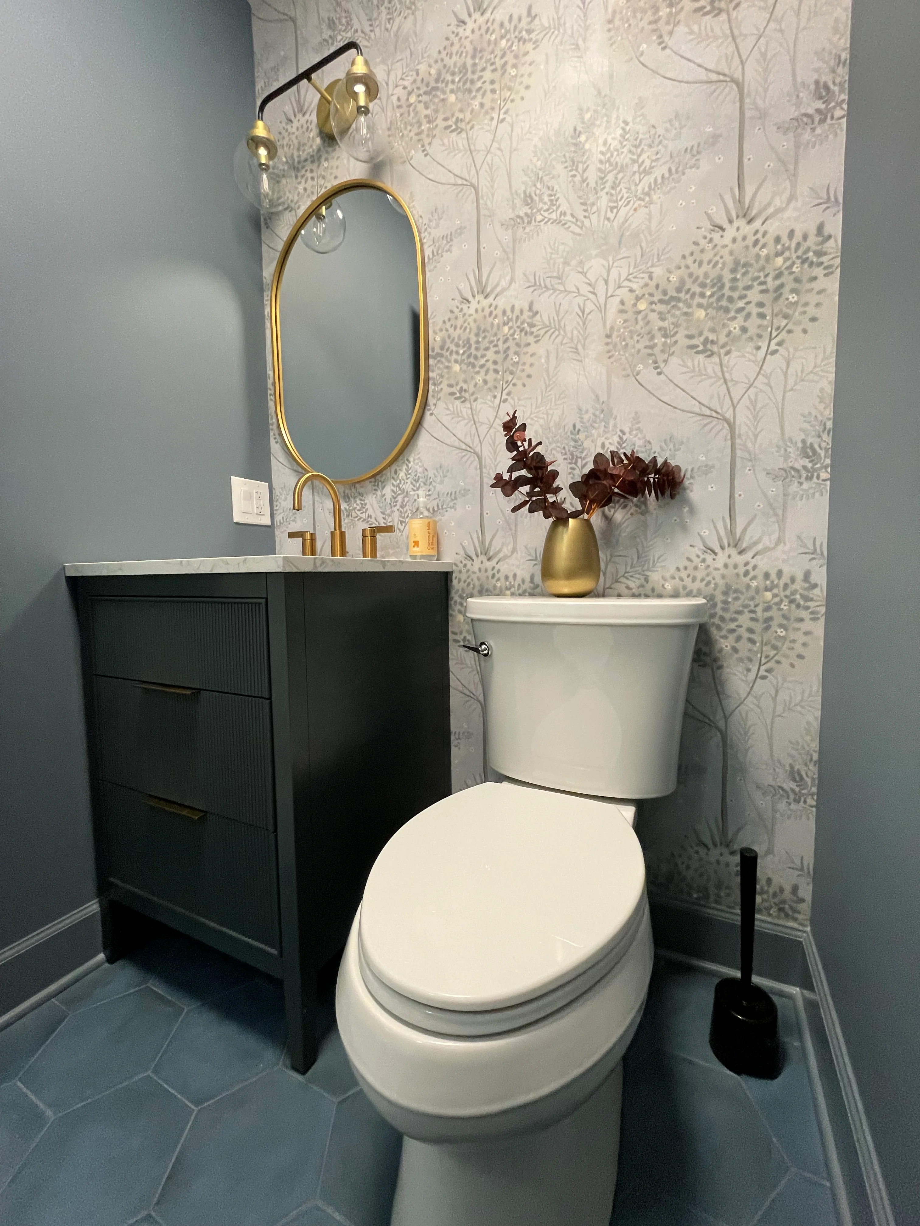 Black vanity with white quartz, brass mirror and fixtures, botanical wallpaper, white toilet, dark tile floor