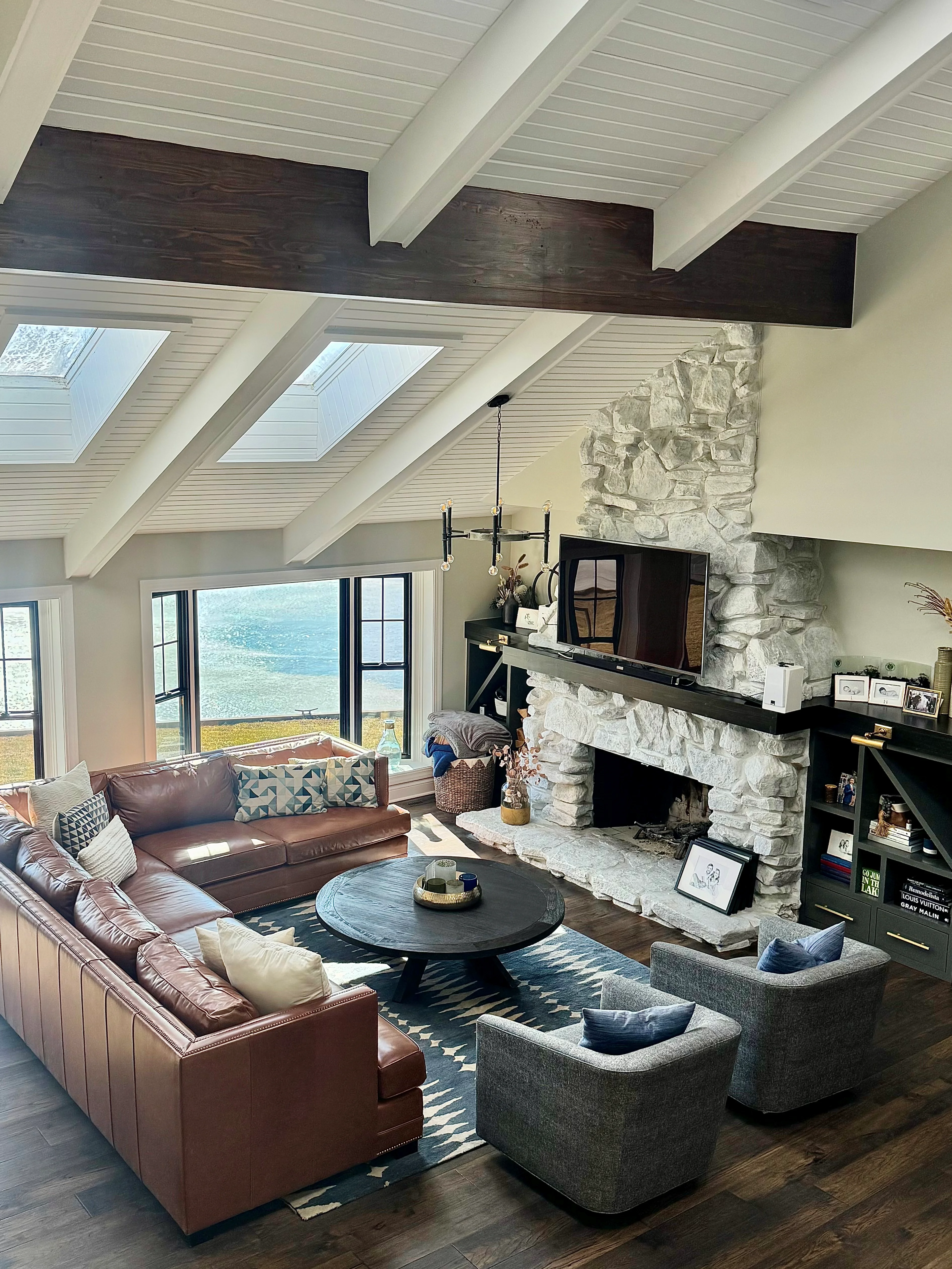Vaulted ceiling with skylights, stacked stone fireplace, dark wood beams, and shiplap soffit