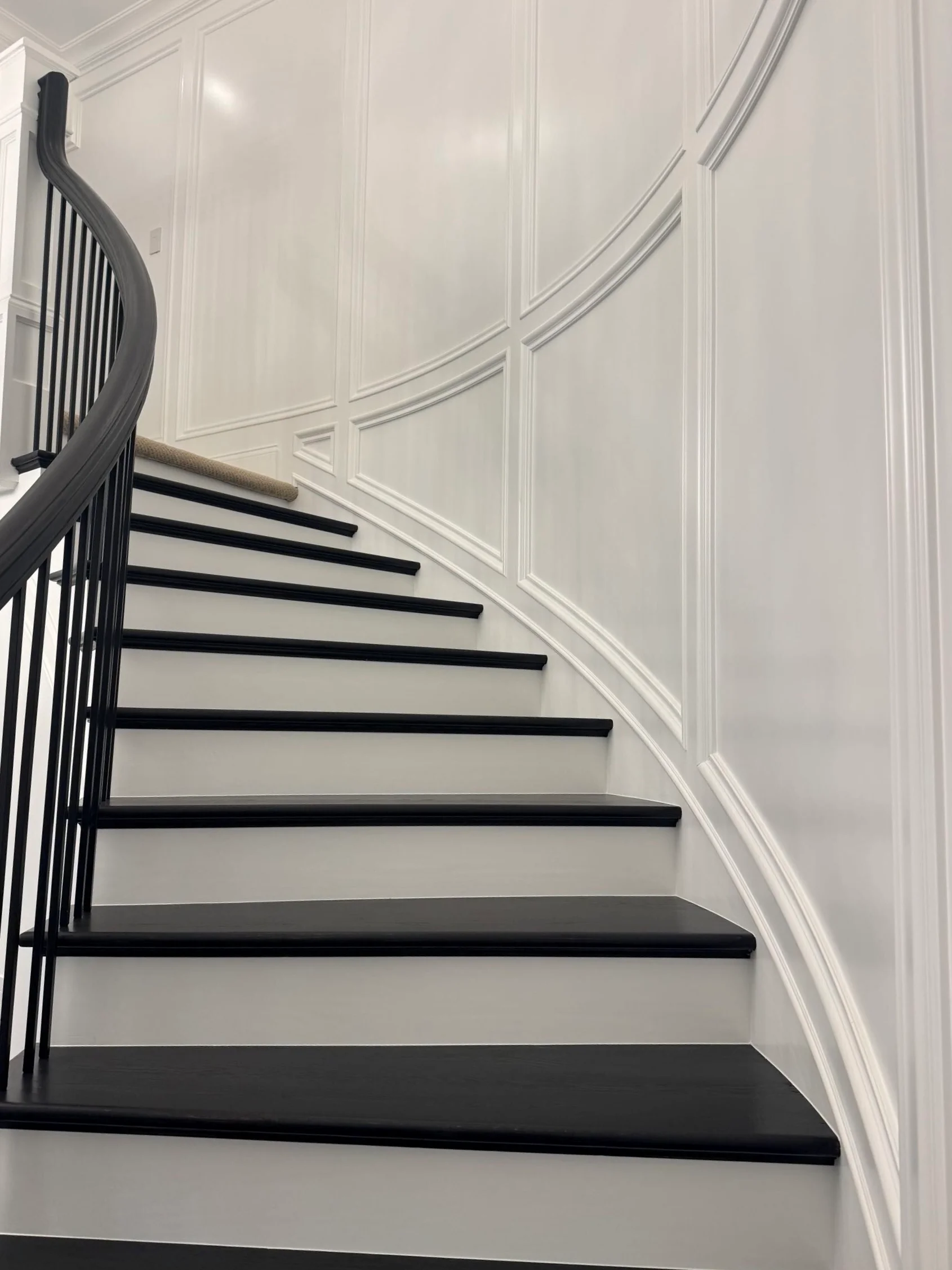 Curved staircase with black spindle railing, gray treads, black risers, and white wainscoting walls