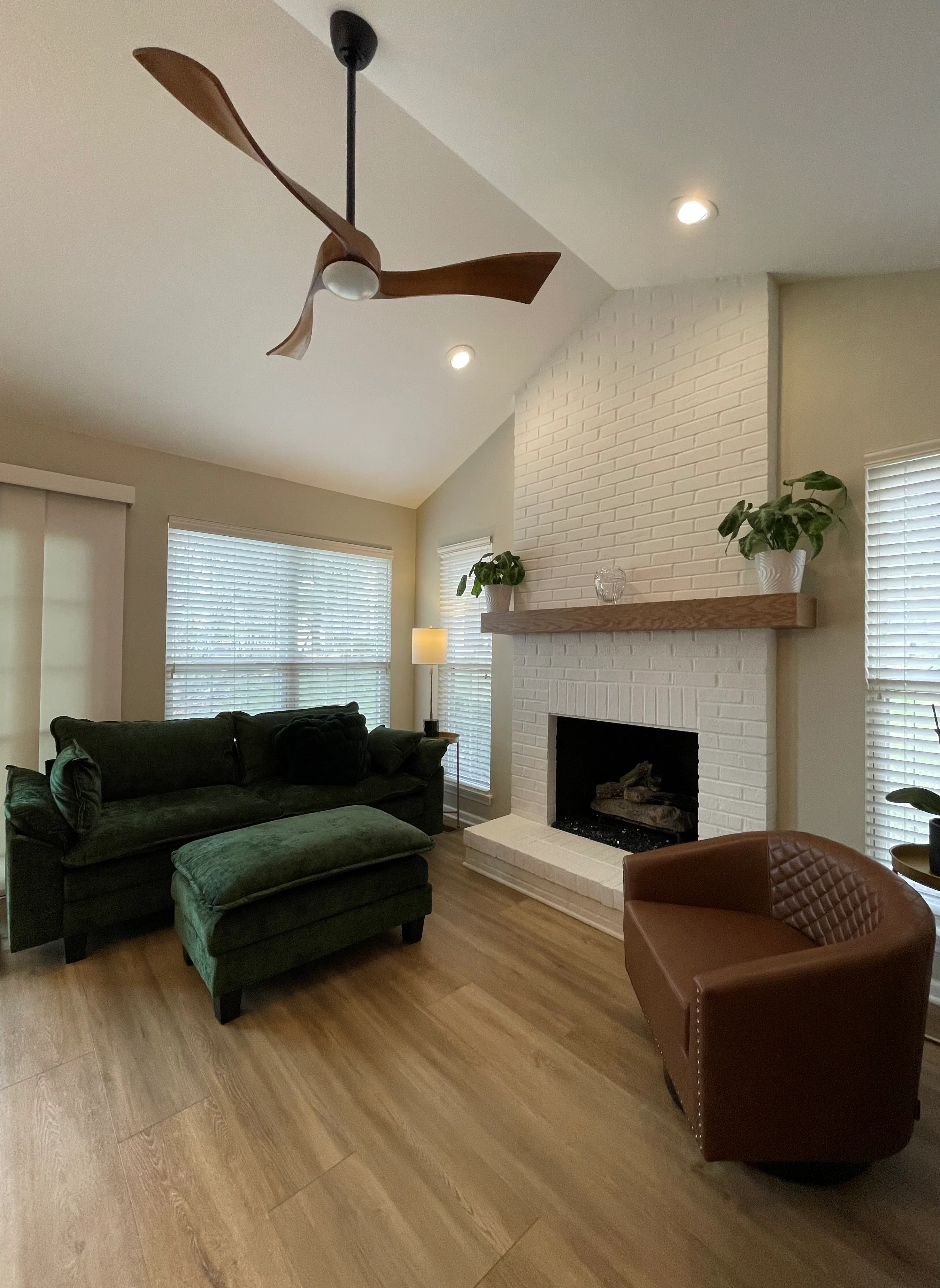 Living room with white brick fireplace, wood ceiling fan, recessed lights, and plank flooring