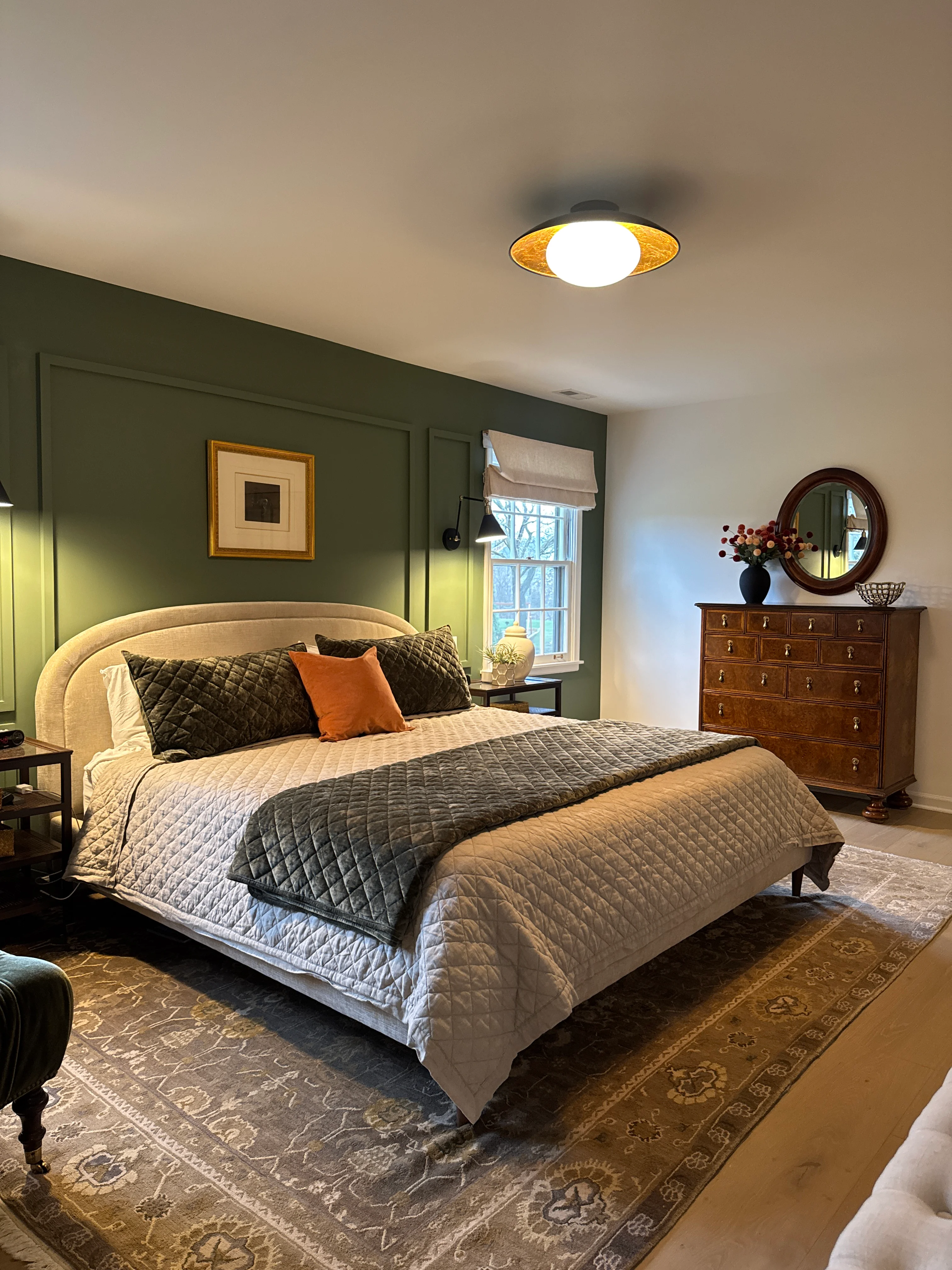 Renovated master bedroom with sage green accent wall, cream upholstered bed, and wood dresser