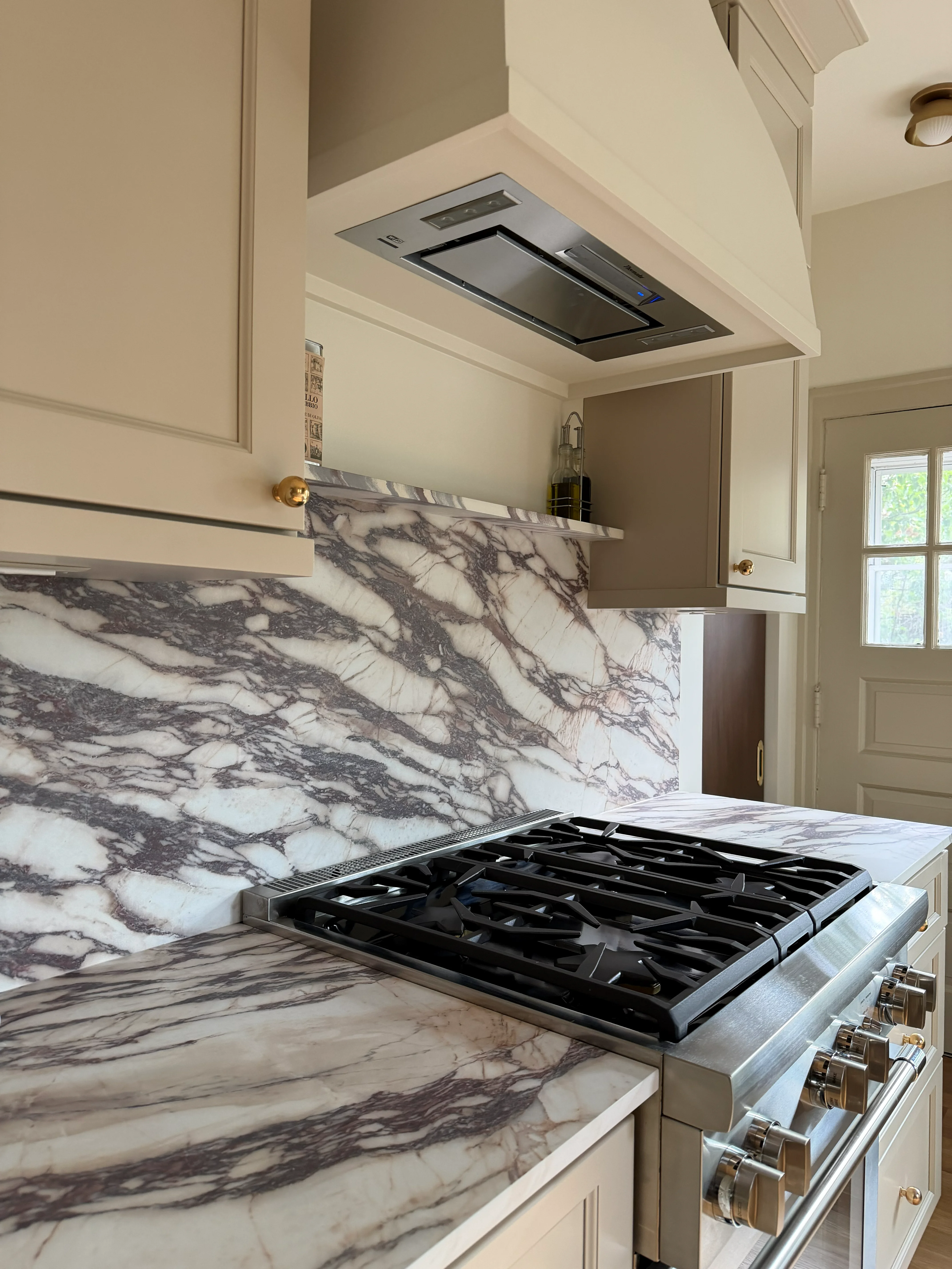 Modern kitchen with white cabinetry, marble backsplash and countertops, stainless steel gas range, and ceiling-mounted hood