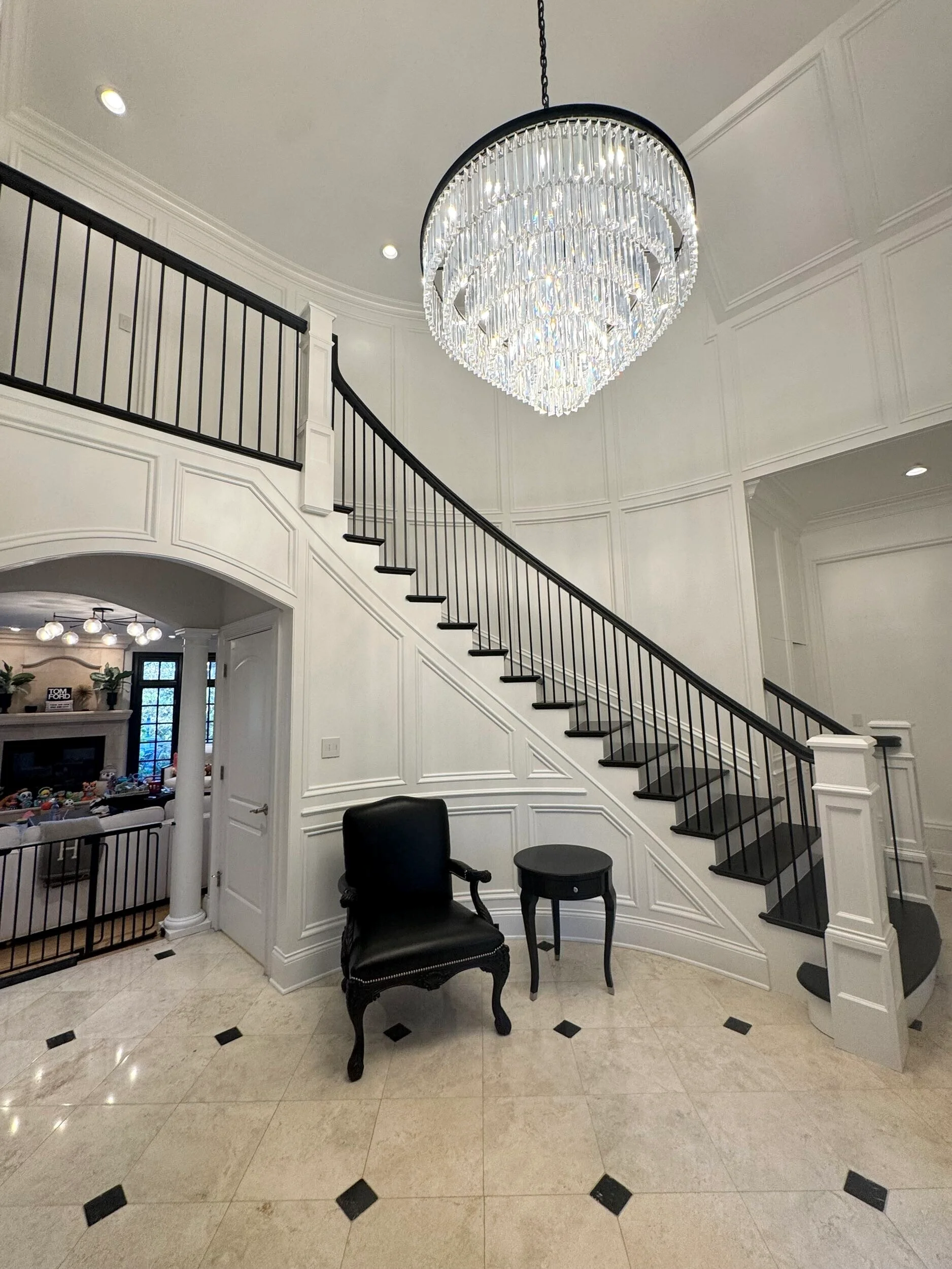 Curved staircase with black balusters, white wainscoting, marble flooring, and crystal chandelier in modern foyer